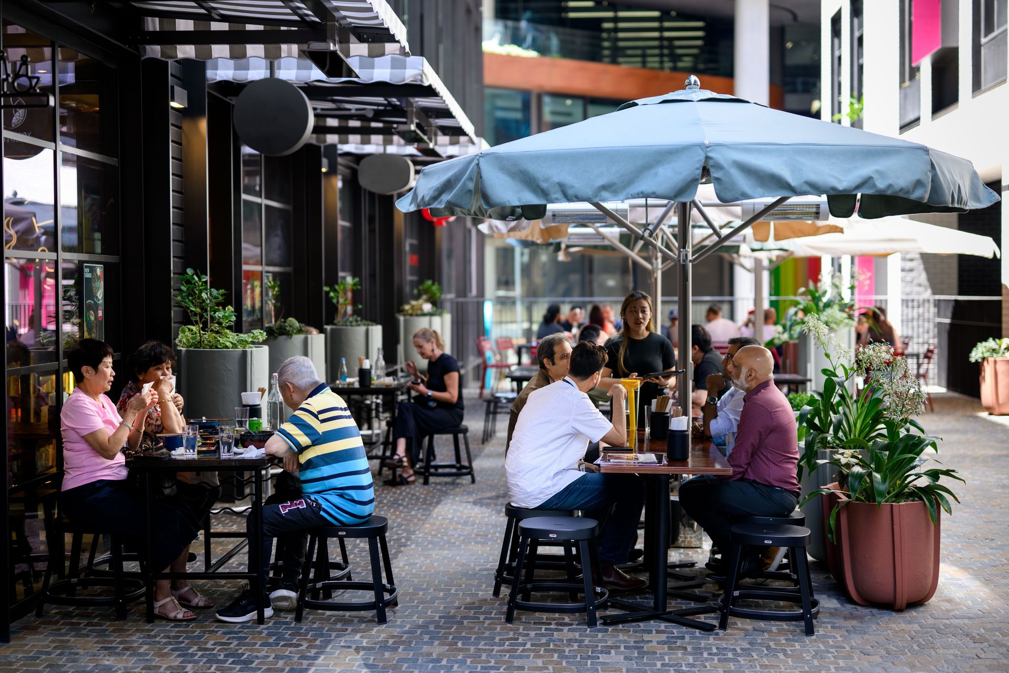 People dining and conversing at outdoor tables under umbrellas, in a stylish urban area with potted plants and contemporary buildings.