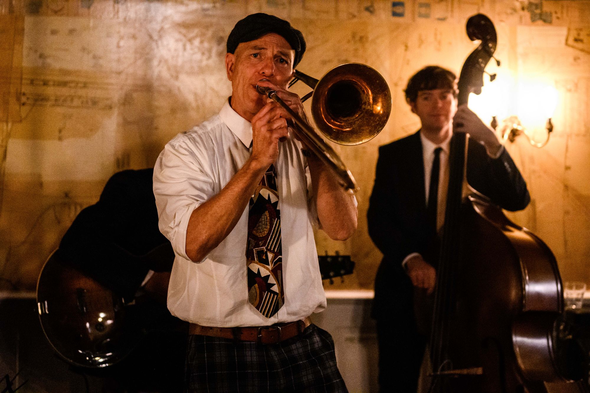 A musician plays a trombone passionately, accompanied by a guitarist and a double bassist, in a dimly lit, cozy jazz setting.