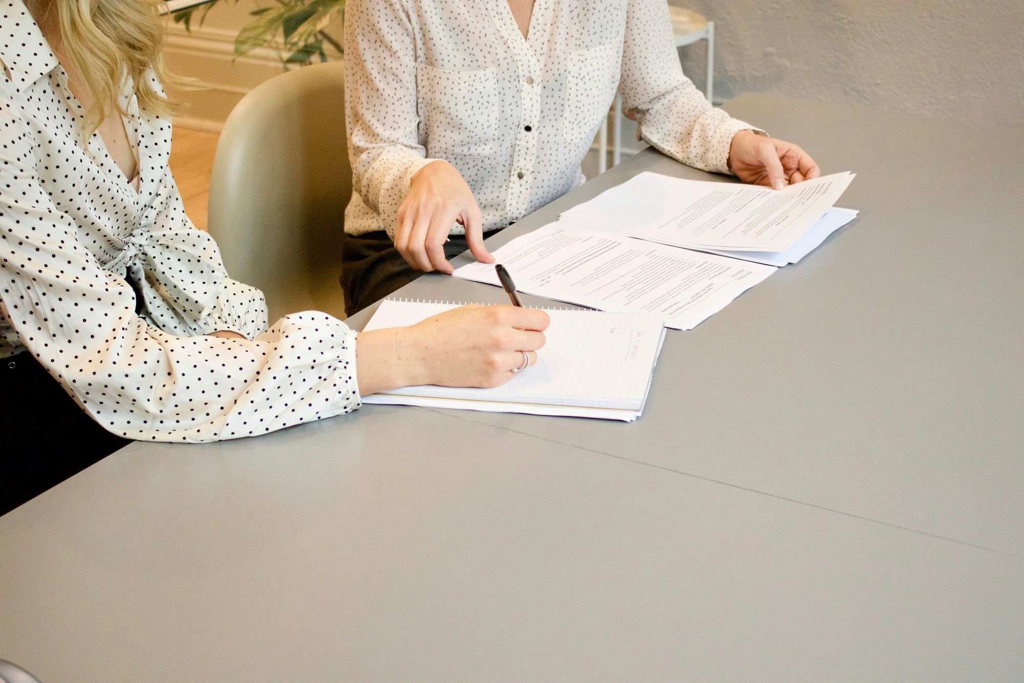 Two people sitting at a desk reviewing documents, one holding a pen. The table is gray, and they both wear polka-dot shirts.