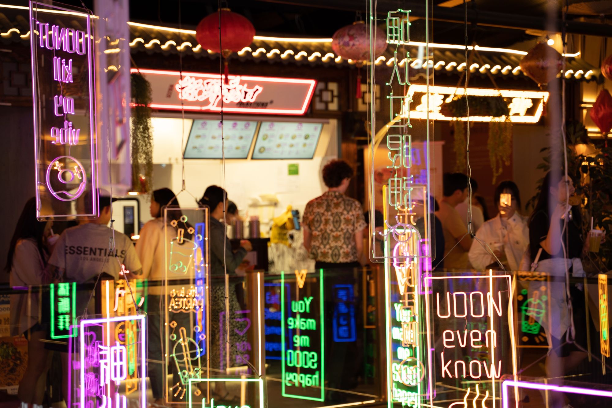 Vibrant neon sign reflections in a bustling restaurant with people inside, featuring various phrases and colorful signage.