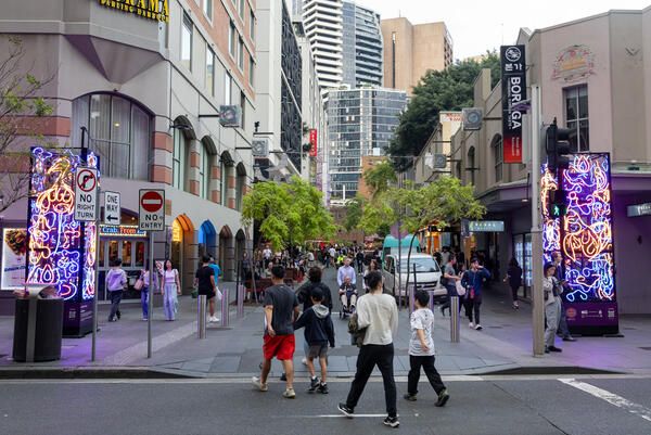 People crossing a city street lined with neon signs and high-rise buildings, featuring trees and bustling shops in the background.