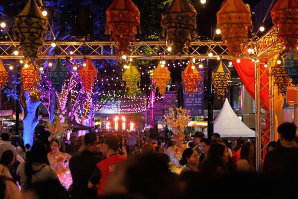 A lively night festival with colourful lanterns, bright string lights, and a crowd enjoying the vibrant atmosphere beneath a canopy of lights.
