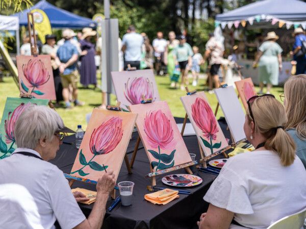 People seated at a table paint pink flowers on canvases during an outdoor art event, surrounded by others in a bustling park setting.