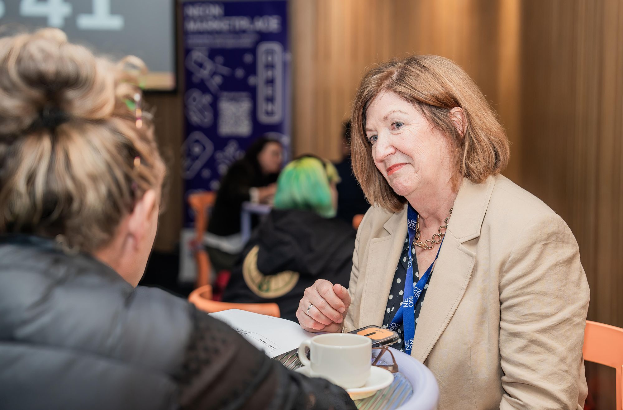 Two women having a conversation at a table with a coffee cup, in a busy indoor setting with people in the background.