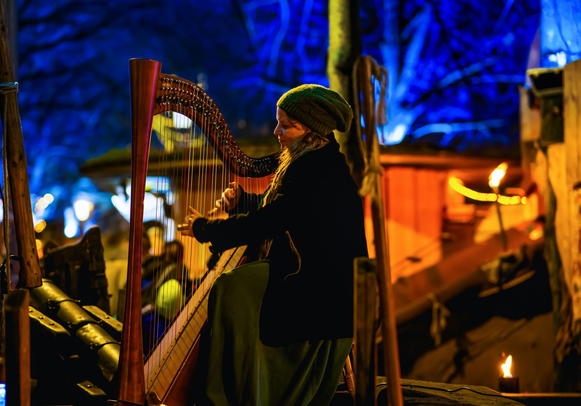 A person plays the harp in a warmly lit outdoor setting at night, surrounded by glowing lights and a cozy atmosphere.