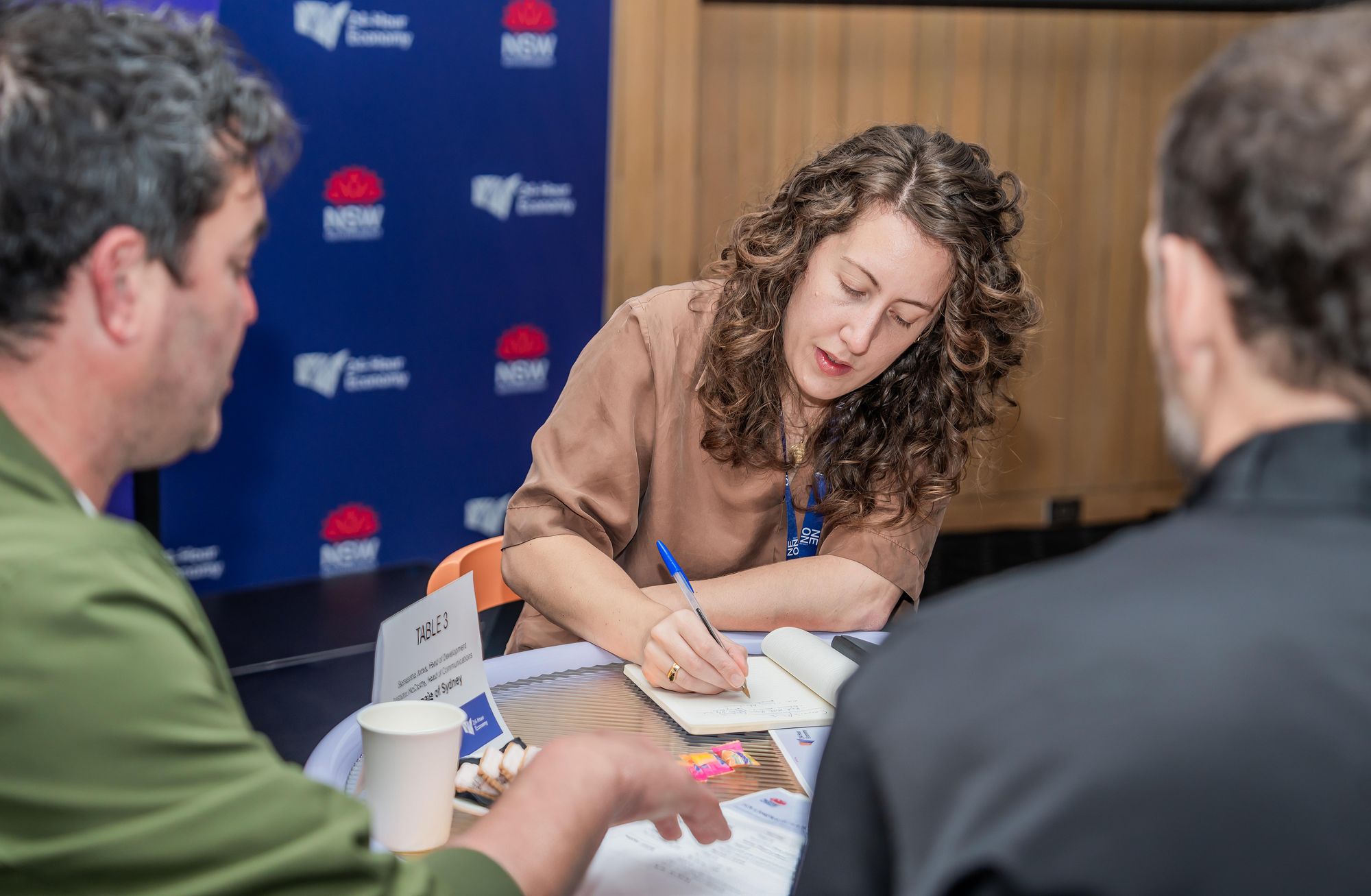 A woman writes notes at a table during a discussion with two men, with a blue event backdrop in the background.