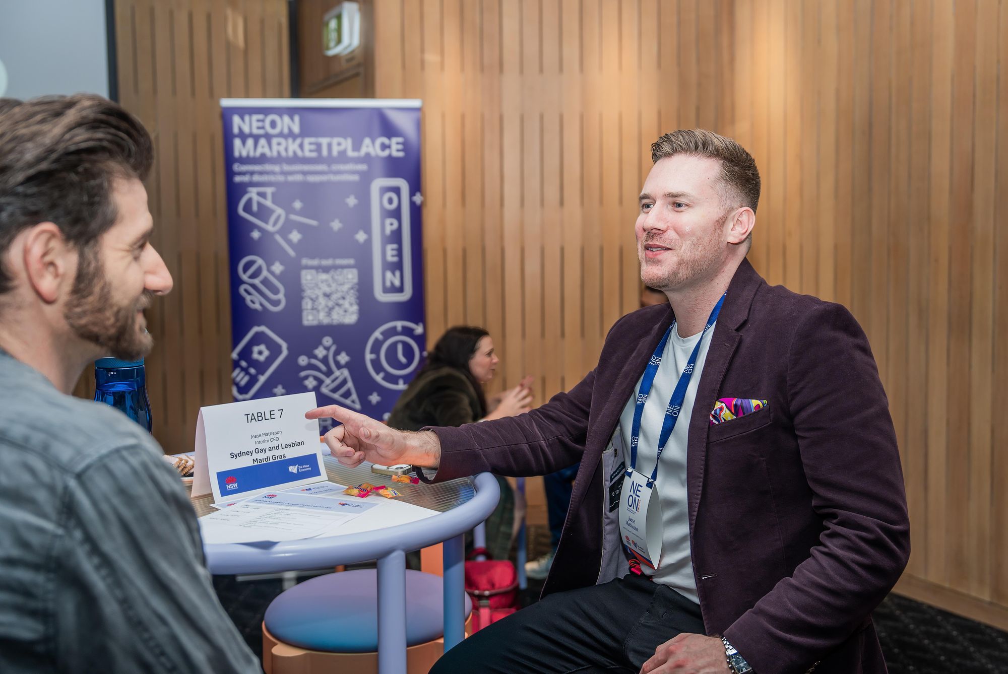 Two men converse at a table in a modern setting. A "Neon Marketplace" banner is visible in the background.