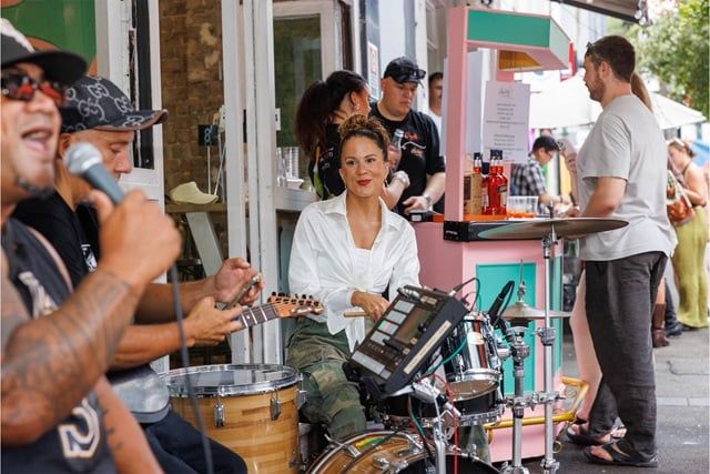 Street musicians perform lively music while people gather near a colorful food stand on a bustling street.