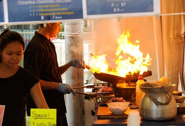 Chef cooking with high flames in a street food stall, while a woman prepares dishes nearby. A "Special" menu sign is visible in the foreground.