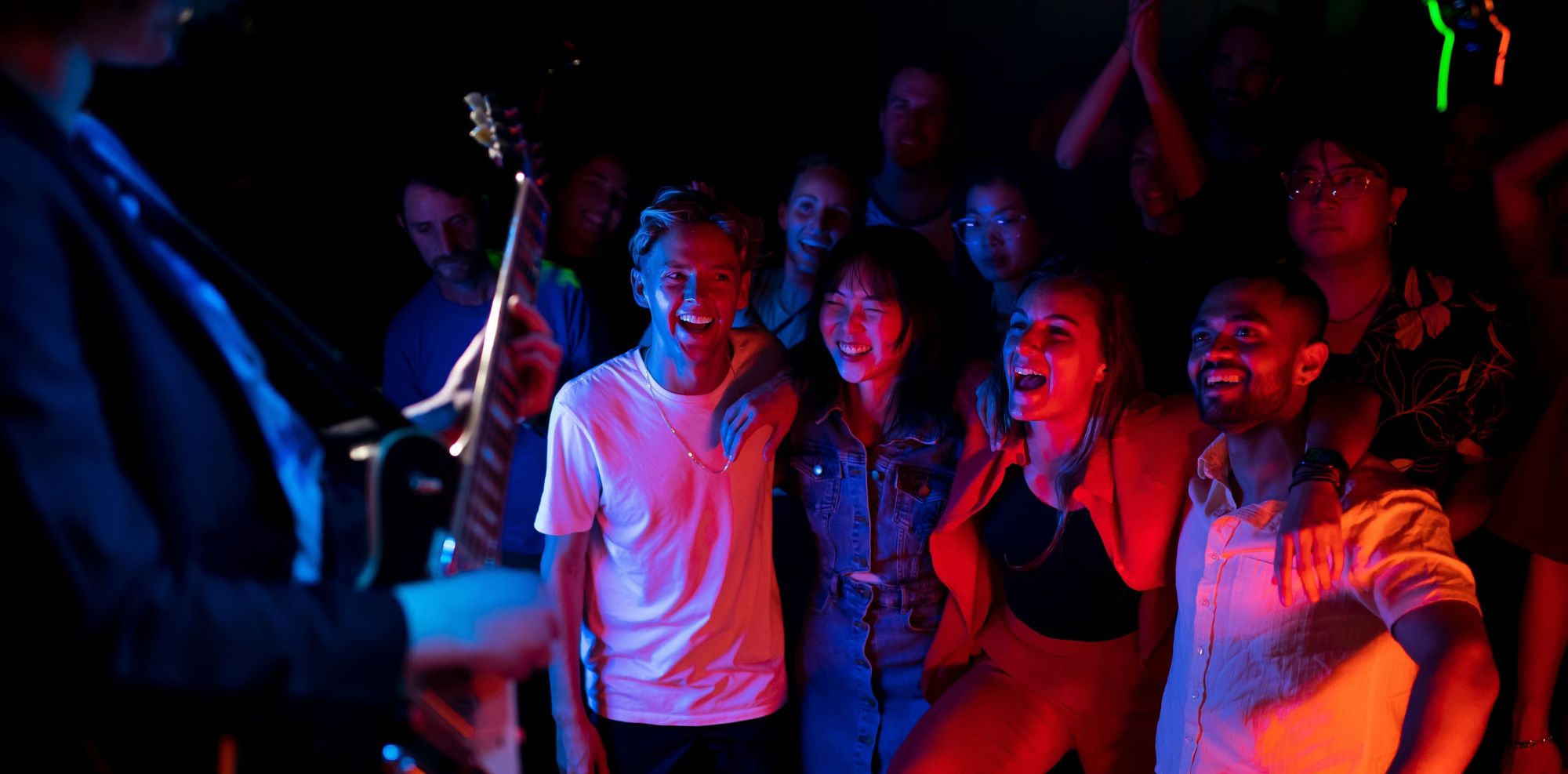 A group of people enjoying a live music performance, smiling and cheering near a guitarist on stage in a dimly lit venue.