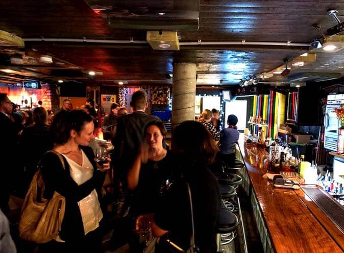 A lively bar scene with people socializing around a wooden counter, colorful walls, and dim lighting.