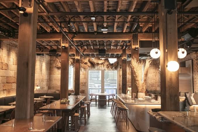 Rustic restaurant interior with exposed wooden beams, stone walls, and hanging lights. Tables and chairs are neatly arranged. Large window at the back.