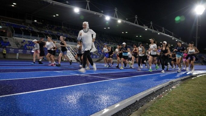 A group of runners starting a race on a blue track at night in a stadium, illuminated by overhead lights.