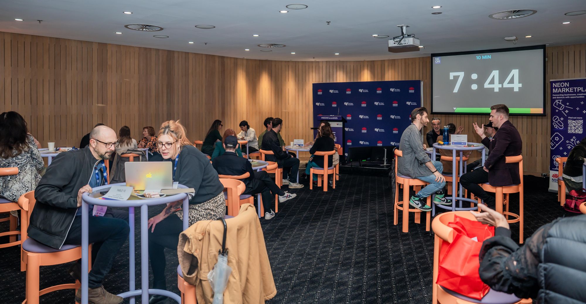 People sitting at tables in a conference room, engaged in discussions. A timer on a screen displays 7:44.
