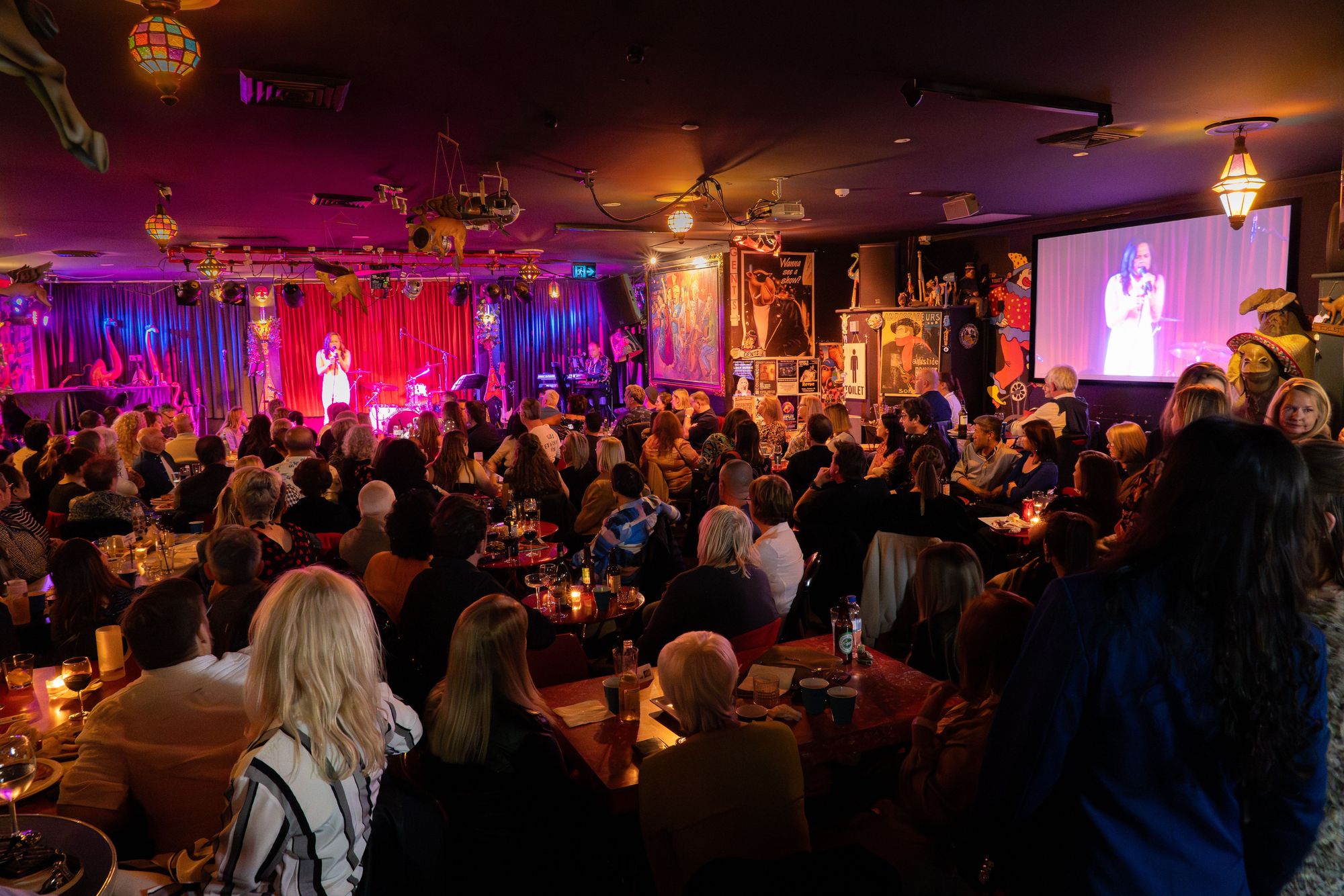 A lively audience watches a performer on stage at a dimly lit venue with vibrant red and blue lighting, as seen from the back of the room.