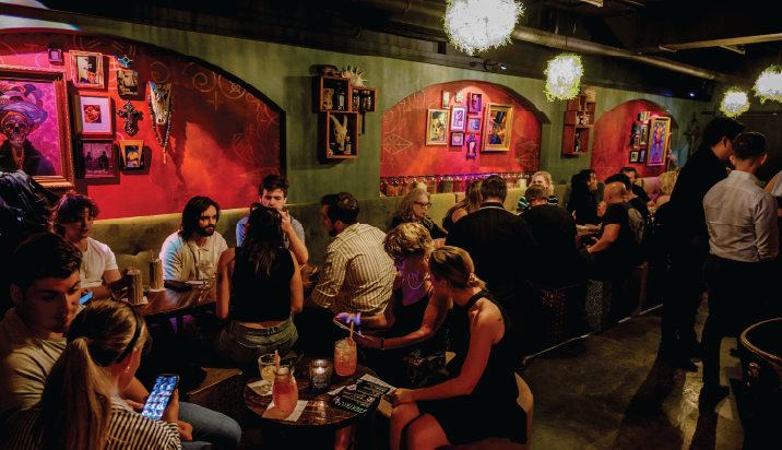 A bustling bar with people seated at tables, chatting and using phones. The walls are decorated with framed art and illuminated by dim, warm lighting.