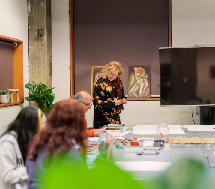 A medium shot of an adult learning workshop or class around a table. A woman with blonde hair wearing a dark patterned dress and scarf stands at the head of the table, looking down at materials. Other participants are seated and partially visible, with a TV screen and two framed artworks visible on the wall behind the instructor.