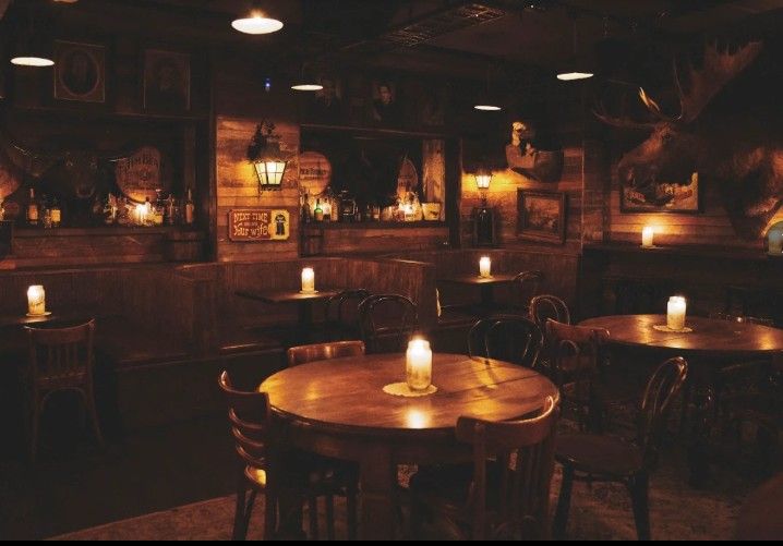 Cozy, dimly lit vintage pub interior with round wooden tables, chairs, wall-mounted decor, and flickering candles creating a warm atmosphere.