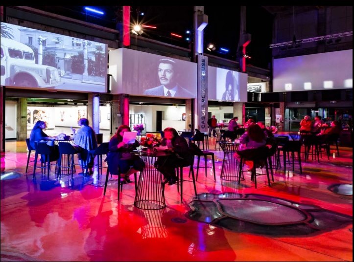 A lively interior shot of a modern event space or hall with a glossy, colorful floor reflecting vibrant red and blue lighting. Guests are seated at small black tables and chairs. Multiple historical film clips and images are projected onto large white walls in the background.