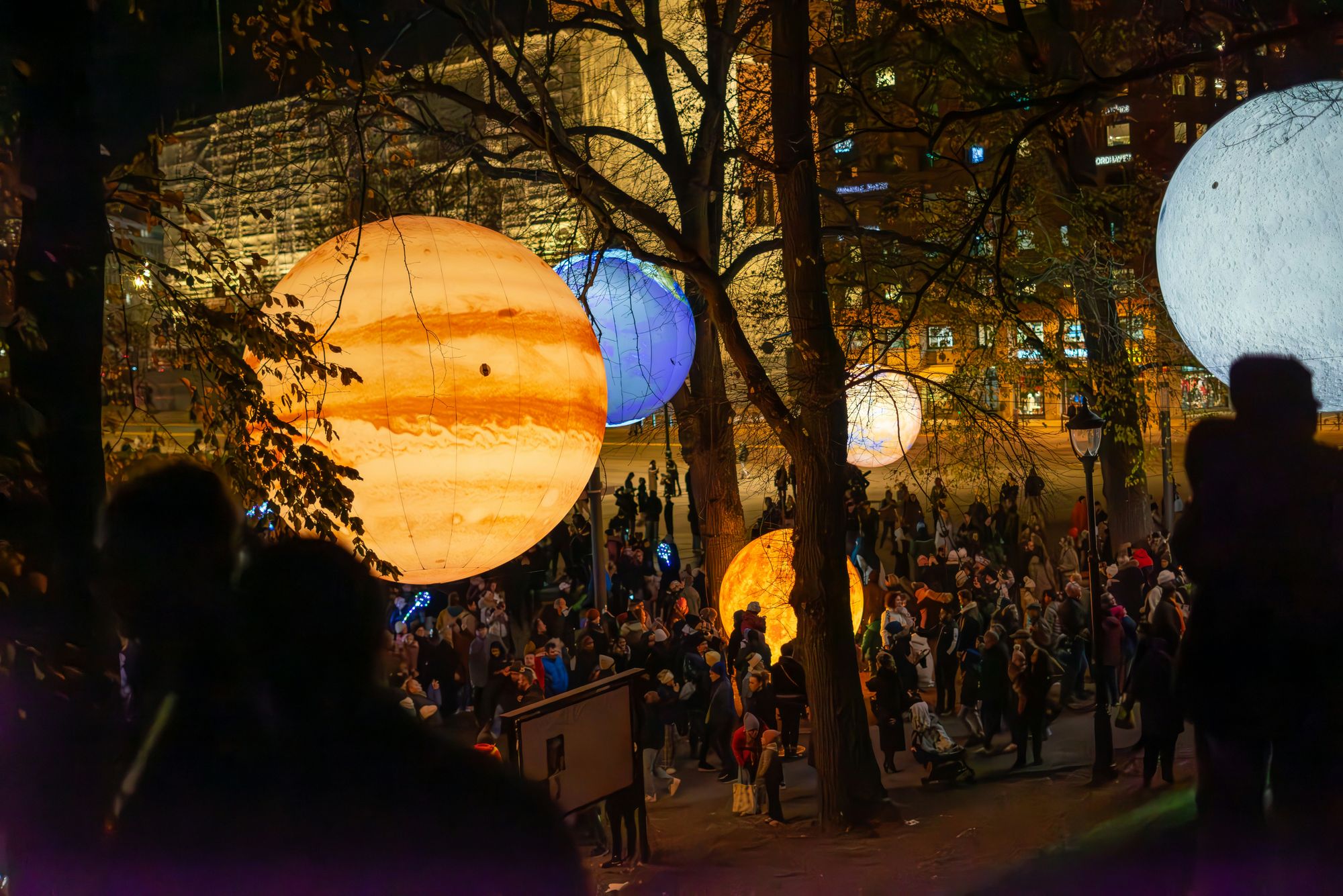Illuminated giant orbs resembling planets hover among trees, as a crowd gathers beneath them during a nighttime outdoor event.