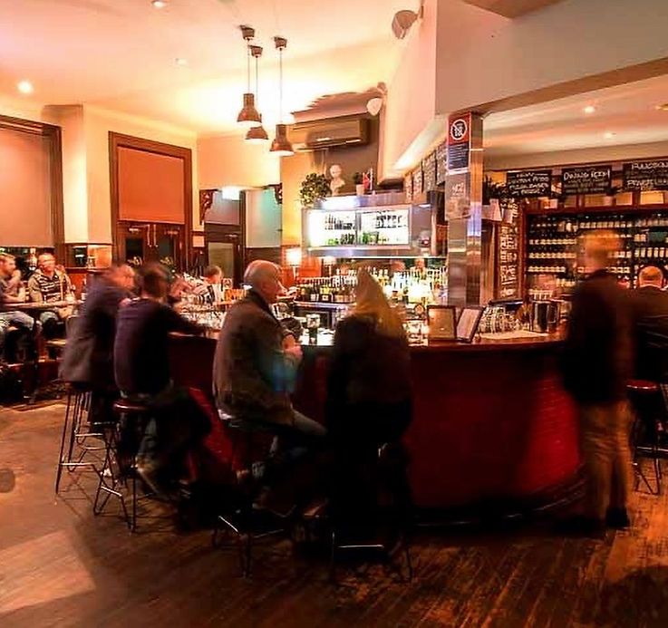People sitting at a warmly lit bar, chatting and drinking. Shelves of bottles line the walls, and a bartender works behind the counter.