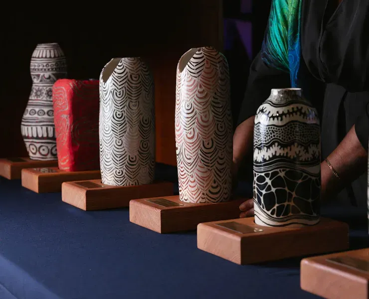 A row of five intricately patterned vases on wooden bases, displayed on a blue tablecloth with a person adjusting one vase.