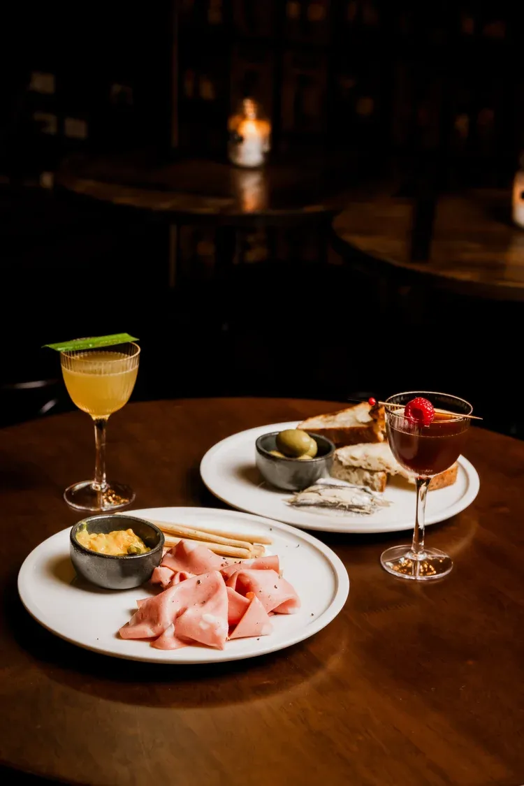 Two cocktails and plates of charcuterie with ham, cheese, and olives on a wooden table in a dimly lit setting.