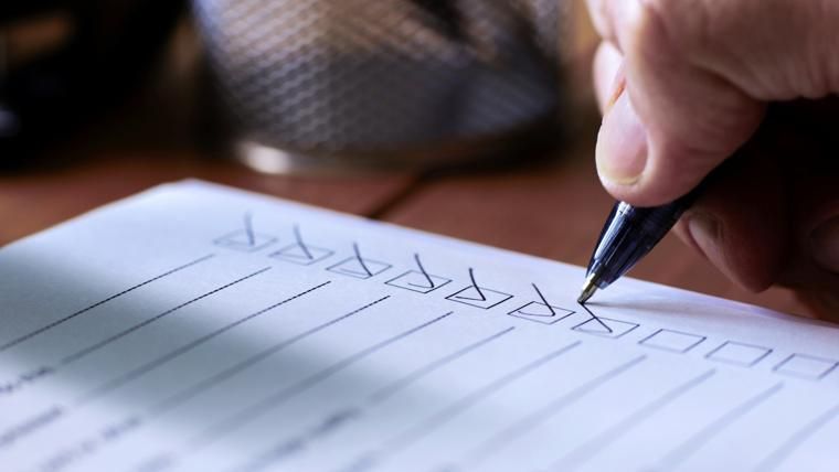 Close-up of a hand using a pen to check boxes on a paper checklist, with a blurred wire mesh holder in the background.