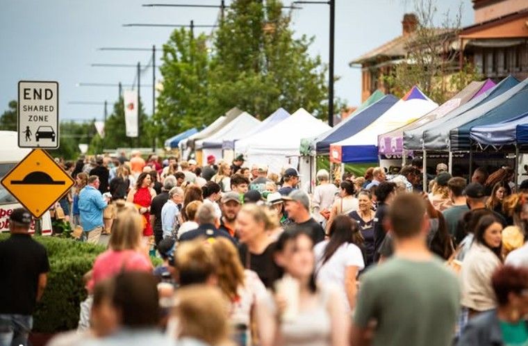 A bustling outdoor market with crowds browsing stalls under colourful awnings, surrounded by trees and buildings. A sign marks the end of a shared zone.