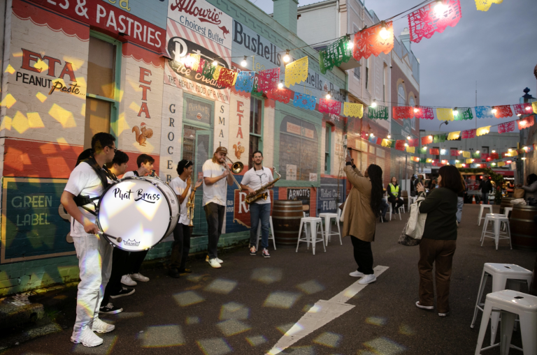 People enjoy live music from a brass band on a lively street decorated with colorful flags and vibrant mural art.