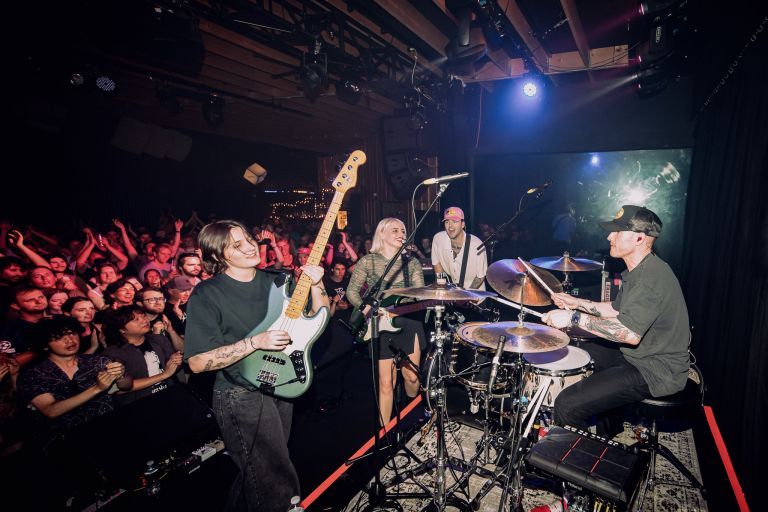 Band performing energetically on stage, guitarist and drummer in focus, with an enthusiastic crowd cheering in the background.