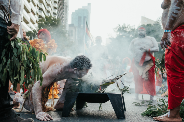 A person participates in a traditional smoking ceremony, surrounded by others in ceremonial attire, on a city street.