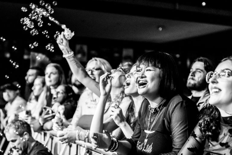 Black-and-white close-up of front-row concertgoers cheering and smiling as bubbles float from a raised bubble wand.