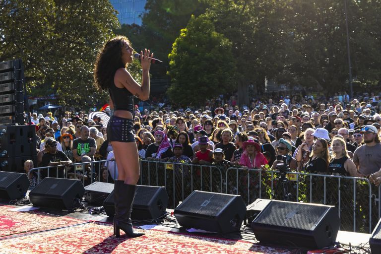 A singer performs on an outdoor stage in front of a large, diverse crowd under sunny skies.