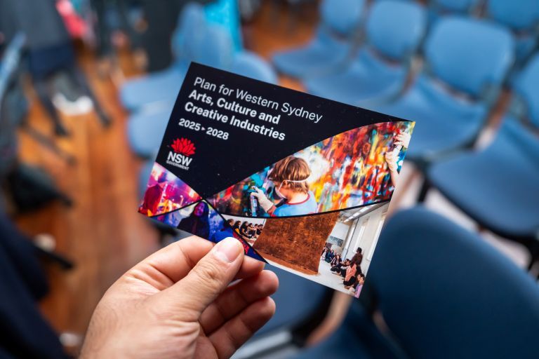 Hand holding a colourful pamphlet titled "Plan for Western Sydney Arts, Culture and Creative Industries 2025–2028" over blurred blue chairs.