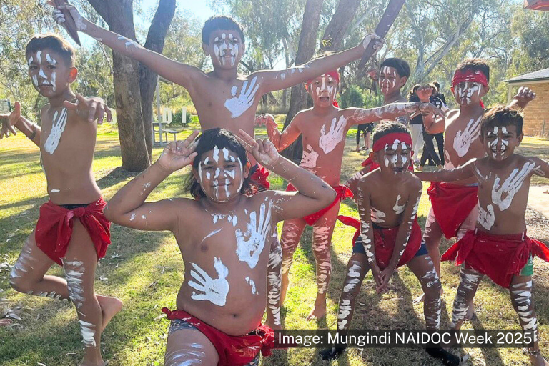 A group of children in traditional attire, with body paint and red cloths, pose energetically outdoors in a wooded area during a cultural event.