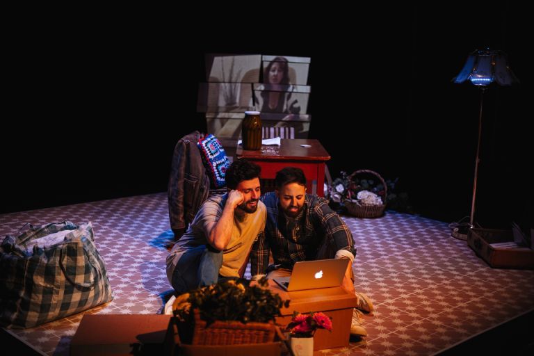 Two people sitting on a patterned floor, looking at a laptop. Behind them are boxes, a lamp, and a projection of a woman's image on the wall.