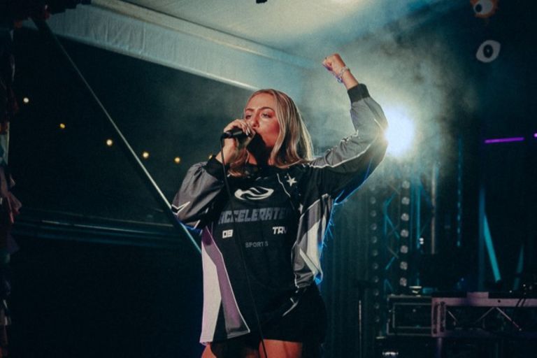 A woman sings passionately into a microphone on stage, wearing a sporty jacket, with her fist raised in the air under dramatic lighting.