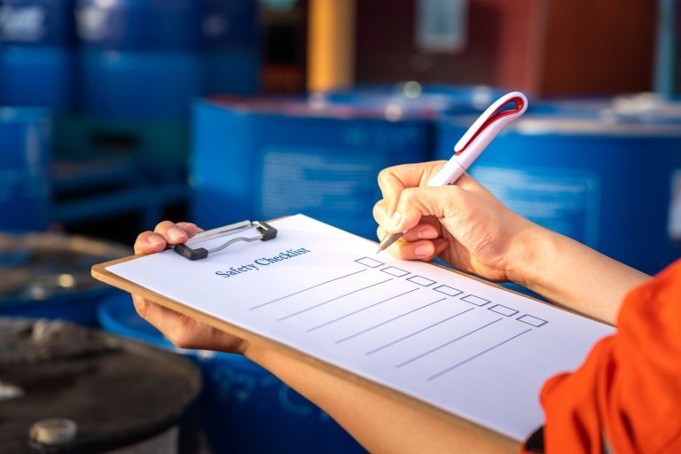 Hands holding clipboard labeled 'Safety Checklist' and marking boxes with a pen in front of blue industrial barrels.