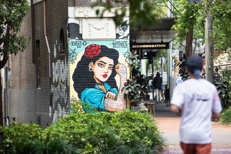 Street art on a brick wall depicts a woman with a red flower in her hair. A person walks by, and a sign reads "Accommodation."