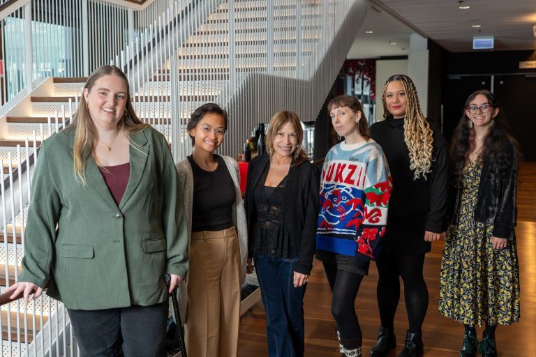 A group of six women standing together in a hallway with a staircase, smiling at the camera.