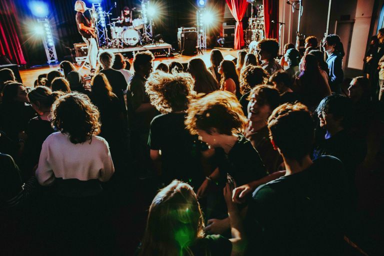 Audience enjoying a live band performance in a dimly lit venue, with colorful stage lights and people dancing enthusiastically.