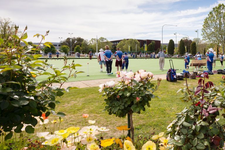 People playing on a lawn bowling green surrounded by colourful flowers, with a clear sky and trees in the background.