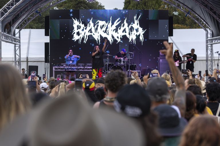 Outdoor concert with band performing on stage under an arched canopy, crowd in foreground and large LED screen with stylized logo behind.