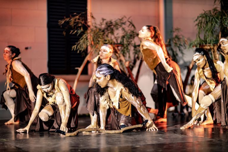 Performers with body paint and traditional attire engage in a dynamic cultural dance on stage, with props and foliage in the background.