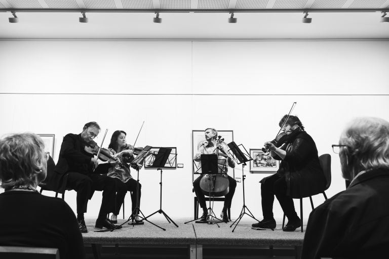 Black and white image of a string quartet performing for an audience in a gallery setting.