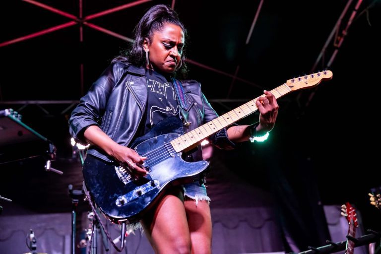 Musician playing an electric guitar on stage, wearing a leather jacket and shorts, under bright stage lights.
