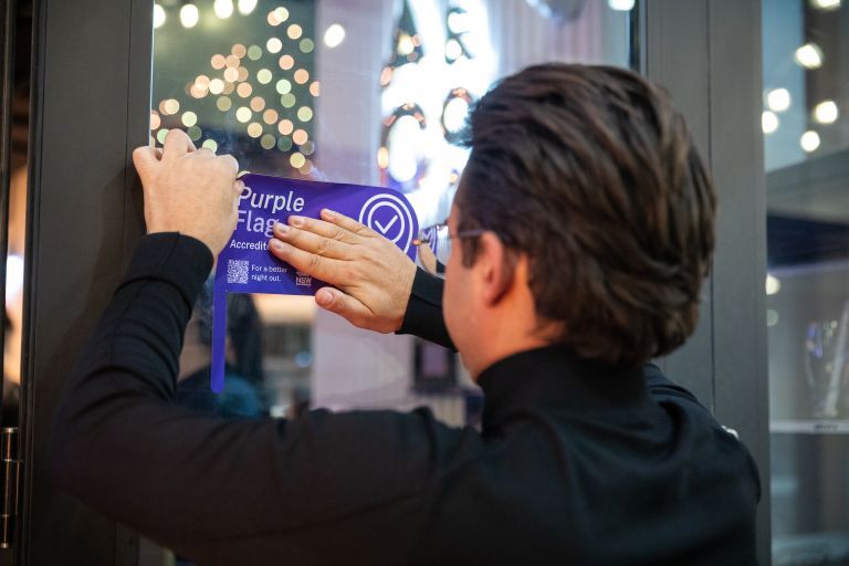 A person in a black shirt applies a "Purple Flag Accredited" sticker on a glass door with blurred lights in the background.