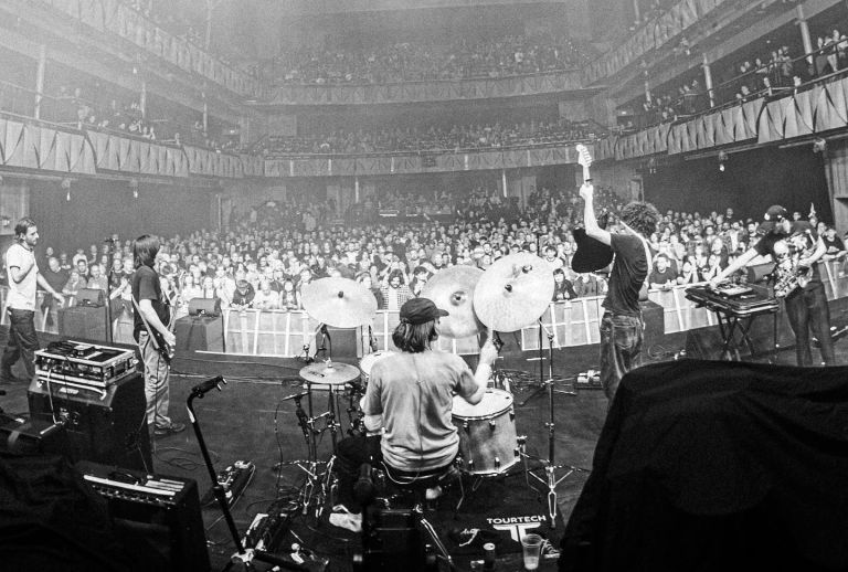 A drummer and musicians perform on stage facing a large audience in a dimly lit concert hall.