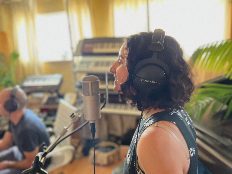 Woman with headphones singing into a microphone in a music studio, surrounded by instruments and soft natural light.